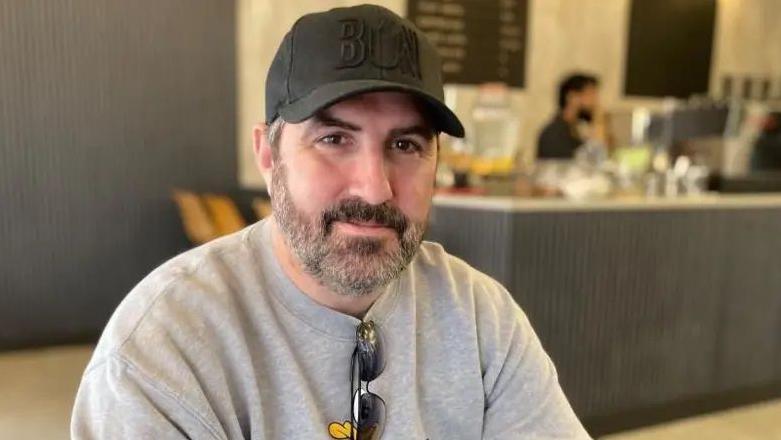 Peter McCormack with a dark beard and wearing a dark green cap and grey T-shirt with sunglasses hanging over the top. He is sitting in a cafe. The serving area can be seen behind with a bearded barista awaiting a customer. A menu board is visible on the wall behind.