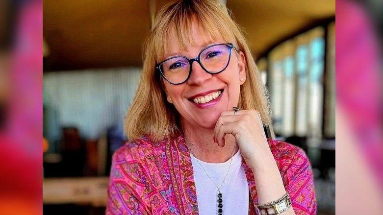 Clare Farthing smiling direct at the camera. She is wearing a bright pink patterned jacket and white t-shirt.