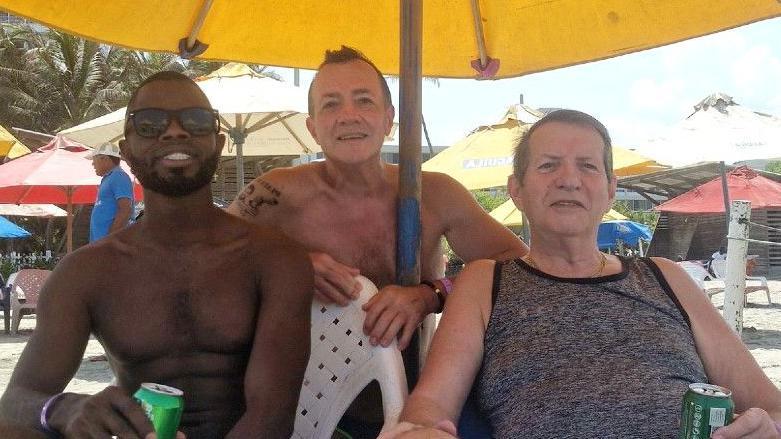 Yostin Mosquera, Albert Alfonso and Paul Longworth are sitting on a beach under a large umbrella, drinking soda and posing for the camera. They are at a holiday resort in Columbia with the sun shining, and umbrellas scattered in the background.