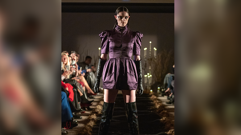 A model on a catwalk wearing a short purple dress surrounded by an audience on either side of her