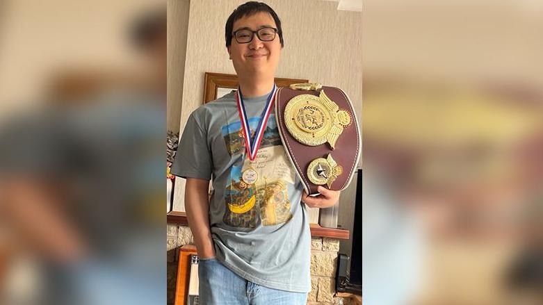 Ha Dang has short black hair and is wearing glasses, a green T-shirt and blue jeans. He is holding the UK winner belt over his left shoulder. It is brown leather with large metal badges on it.