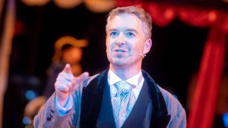 A man is standing on stage wearing a suit. He is pointing towards the camera.