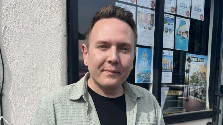 A man with short hair stands in front of a window which has several posters displayed in it. He is looking into the camera and wearing a light shirt with a black t-shirt underneath.