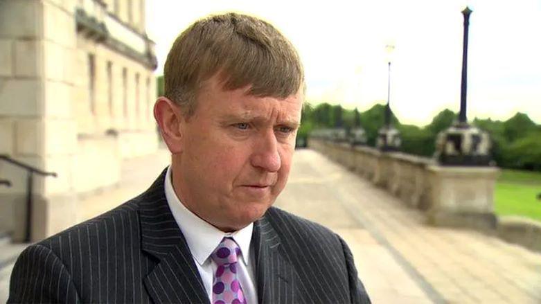 Mervyn Storey standing on the steps of parliament buildings at Stormont wearing a suit and tie.