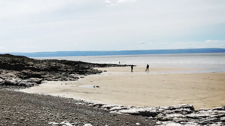 Two individuals are seen standing on a large beach. There are rocks to the left and at the front of the picture. 