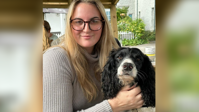 Holly Crouch, a young woman with long, wavy blonde hair and tortoiseshell glasses, is wearing a grey jumper with a high neck as she holds her dog, a black and white spaniel, and smiles at the camera.