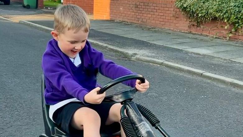 A very excited and happy-looking young blond boy in his purple school uniform jumper, white shirt and dark shorts rides a go-kart in the street.