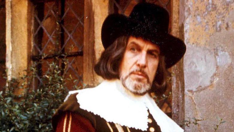 A slightly blurry image of Vincent Price (1911-1993), playing Matthew Hopkins. He is wearing a black wide-brimmed hat, a maroon jacket with gold buttons and braiding and over it a broad lace-trimmed collar. He is standing against a stone wall and mullioned window.