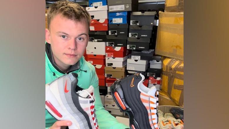 A young man with short blonde hair wearing a green jacket and crouching down in a warehouse holding two Nike trainers for show. with shoe boxes in the background