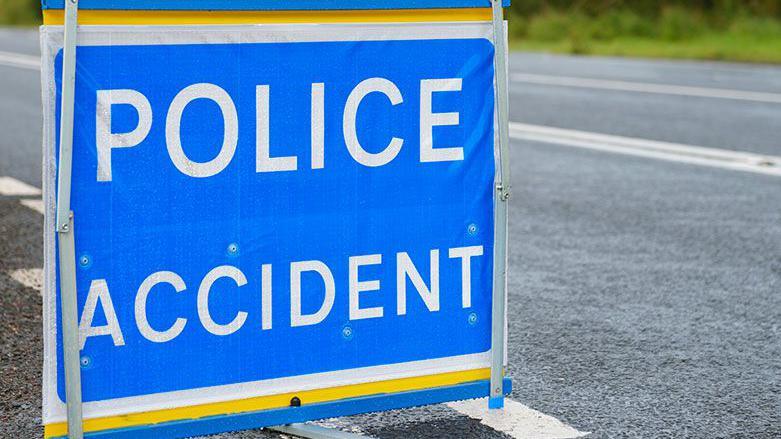 Police accident sign, white bold letters on blue, on a road, with grass in background