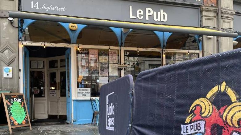 The outside of Le Pub, which has a grey sign with white writing. There is also  a sign outside that says "Le Pub is for lovers"  