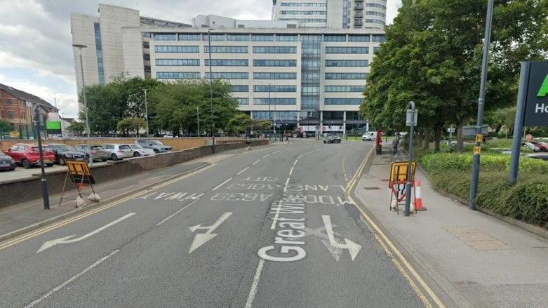 A city centre road. To the right is a sign marked 'Asda Home Office' and to the left is a car park. In the middle is a three-lane road with markings indicating it is one-way. A block of flats stands in the distance.