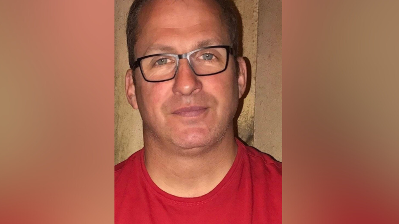 A middle-aged man stands wearing a red-shirt with the word BOSS on the front and black framed glasses and a neutral expression. 