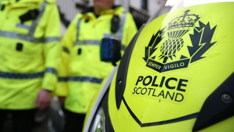 Generic image - close up of the front of a Police Scotland motorbike with the logo in black and two police officers wearing high visibility jackets standing in the background out of focus