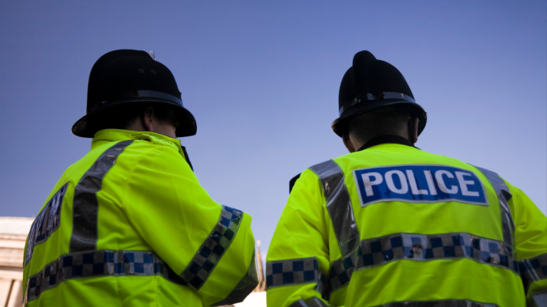 The back of two uniformed police officers wearing hi-vis jackets and black police hats, underneath a bright blue sky