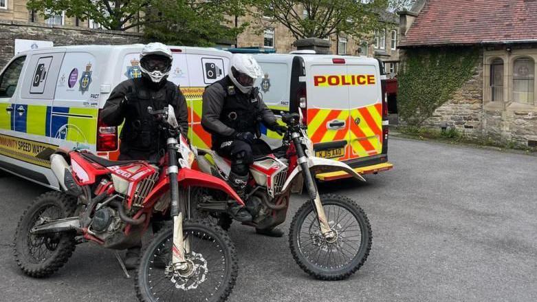 Two police officers in helmets on motorbikes. There are two police vans in the background. 