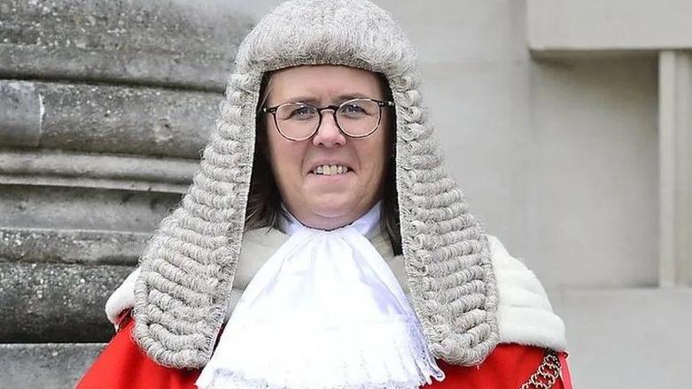 A woman stands in front of a stone building wearing a red robe, a gold chain  and a wig