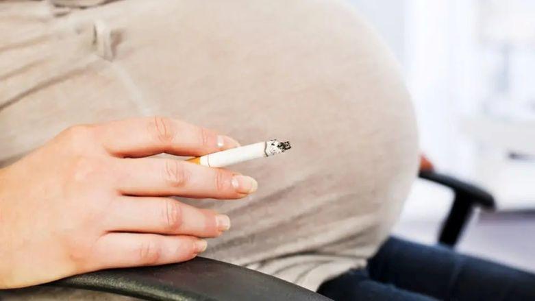 A close up of a pregnant woman's belly. She is sat in a black chair and is wearing a beige top and blue jeans and is holding a cigarette in her hand.