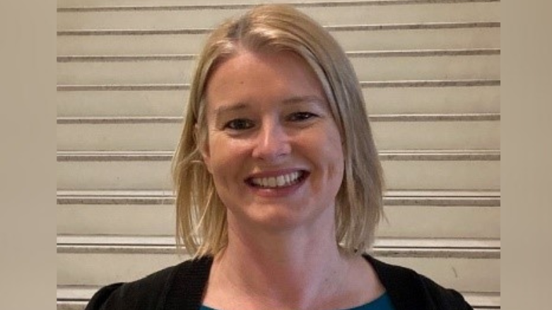 Anne Madden - a woman with shoulder-length blonde hair smiles at the camera. She is wearing a black cardigan and is standing in front of white stairs.