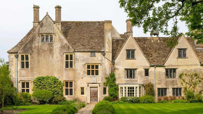 The front of a 16th Century manor house - lots of pitched roofs, stone window frames and chimneys. A large neat lawn and pathway out front, with tree branches hanging over from the right
