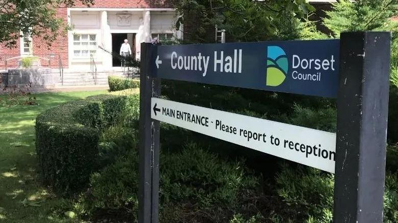 A sign points to the main entrance of County Hall in Dorchester, across a grassed area. The Dorset Council HQ in the background is a brick building with white steps leading to a large doorway.