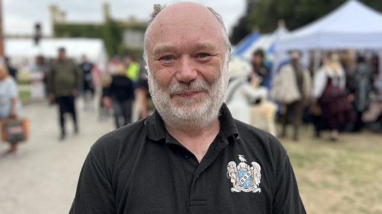 John Naylor, who runs the event, wearing a black v-neck t-shirt with a coat of arms symbol on it. He is stood in the middle of the festival with people milling around in the background and he has a grey beard and grey hair. 