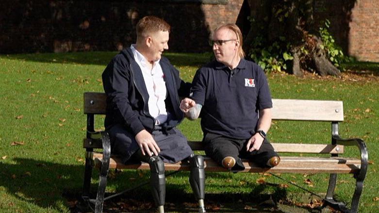 David Jack Plant and Antony “Coops” Cooper sit on a bench in the sunshine, on a grassy patch of land. They are looking at each other. They are both missing their legs, and one has prosthetics