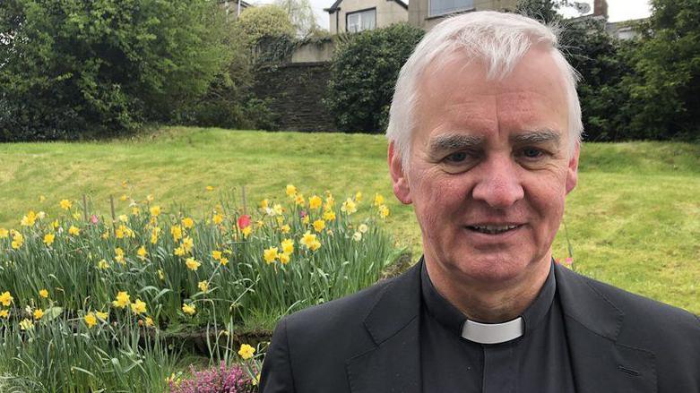 Fr Michael Canny in clerical dress stands in front of a garden of colourful flowers and grass