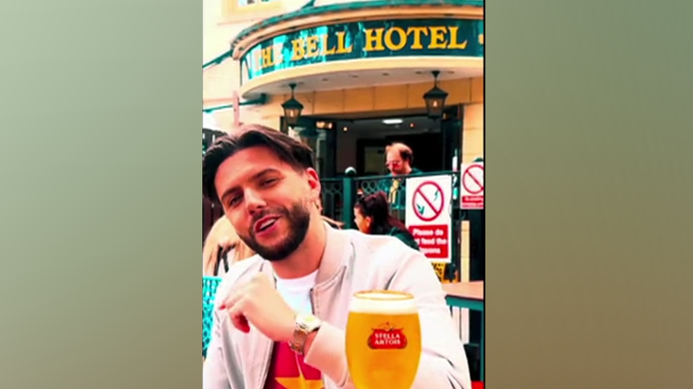 Man smiling. He has a beard and a glass of lager in front of him. He is outside a pub.