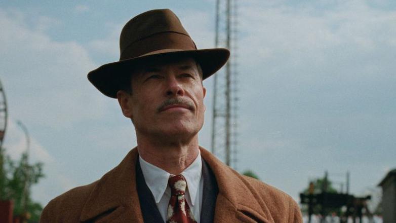 Guy Pearce in a still from The Brutalist. He is outdoors, wearing a smart brown coat and hat, with a shirt and tie.