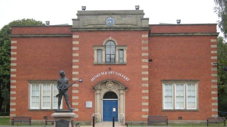 Nuneaton Museum & Art Gallery, a brick-built building with a statue of a solider outside. There is a set of blue double doors, windows on both sides of the ground floor and one window on the upper floor, plus a blue clock on the ornamental features on the top of the building.