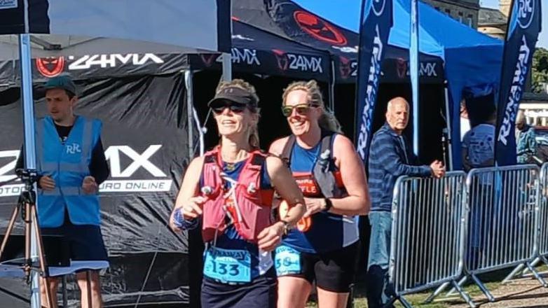 Mrs Hayward-Brackenbury is pictured running a race. She is running with a friend who is slightly behind her. Mrs Hayward-Brackenbury  is wearing a blue vest with a number on it, with a red vest over the top that carries water. She is wearing a black visor cap and black sunglasses.