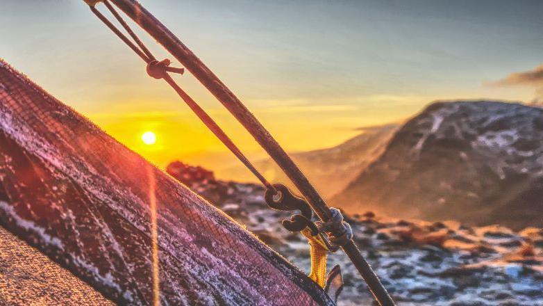 A frosty tent at the top of a mountain range with a yellow/orange sunrise in the distance, next to snow-covered peaks.