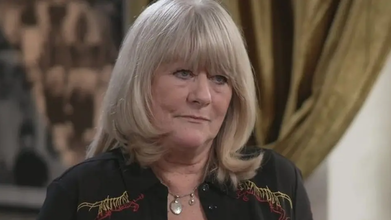 Christine Flack looks to the right of the camera as she is being interviewed. She has a solemn expression and is wearing a dark blouse. She has long blonde hair with a fringe.