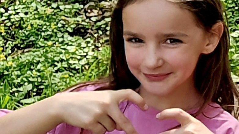 Aria Thorpe. She is pictured outside in front of a hedge. She has long dark hair and brown eyes and is looking ahead, making a heart symbol with her fingers.