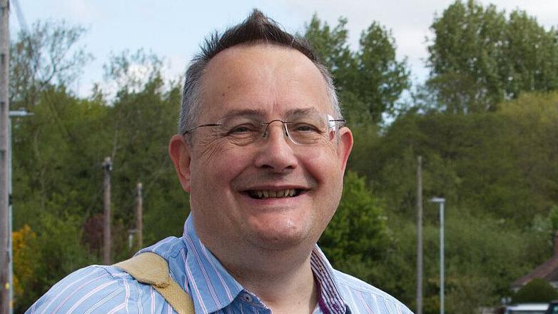 Councillor Paul Semple wears a blue shirt and glasses and smiles at the camera. Lamposts and trees are in the background.