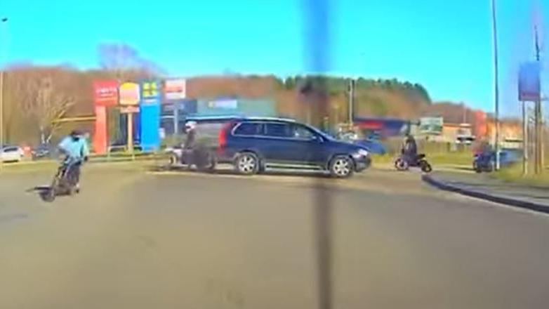 A still from dashcam footage showing a black car blocking a road on a roundabout and at least four people on bikes around it