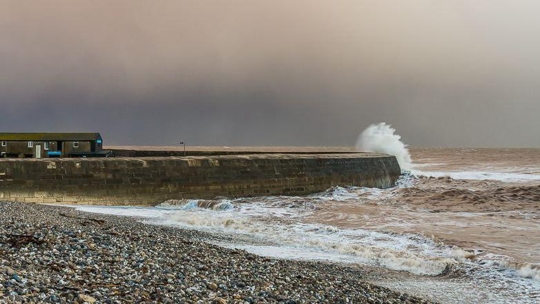 View from the shingle beach of the seaward side of the Cobb breakwater. It is a stormy-looking sky and the sea is crashing against the wall. On the left on top of the wall are two buildings with pitched roofs and chimneys.