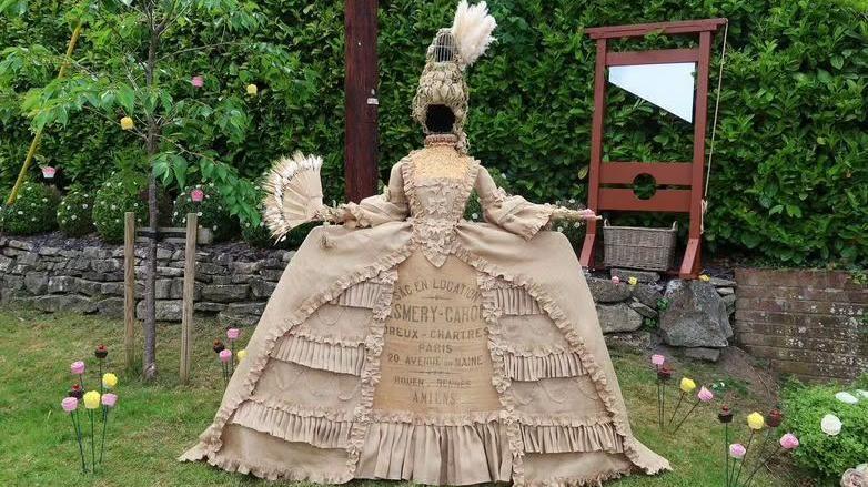 A scarecrow of Marie Antoinette. It is dressed in period costume consisting of a wide, beige dress and a tall blonde wig. 