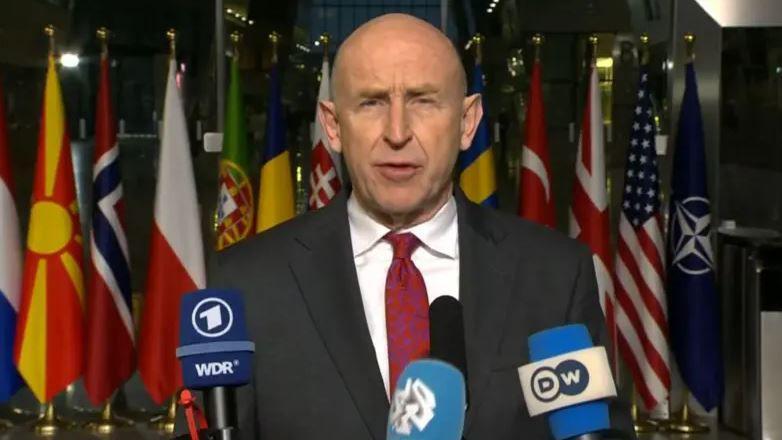 John Healey, who is bald, wears a black suit with a white shirt and red tie as he addresses a NATO meeting on Wednesday