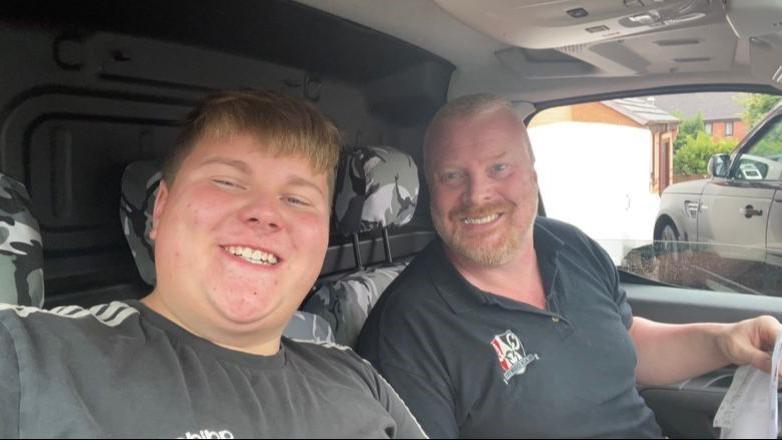 A teenager and a man sit in the front of a vehicle, smiling at the camera. The teenager sits closest to the camera and is extending his arm to take the photo of them.