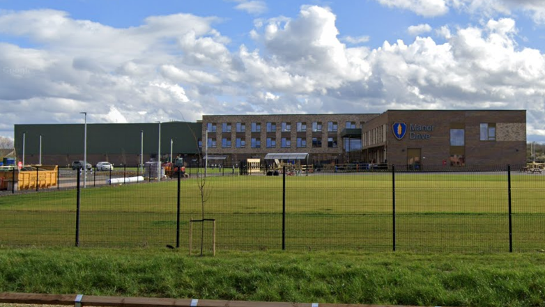 A large brown bricked building is behind a green fence. The building has multiple windows and a sign which says Manor Drive.