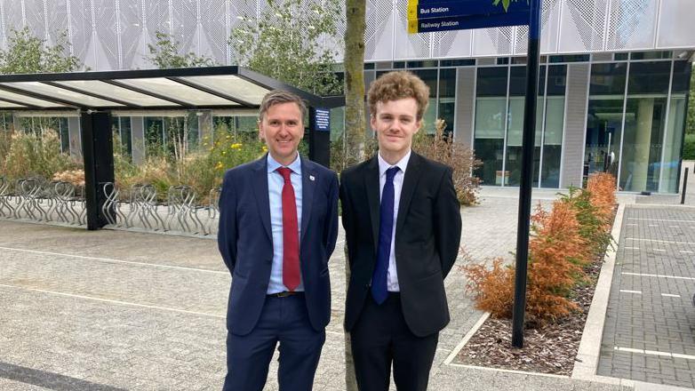 Andrew Pakes and Sam Carling face the camera with their hands behind their backs. They both wear dark coloured suits and ties. In the background is a cycle park and glass fronted building.