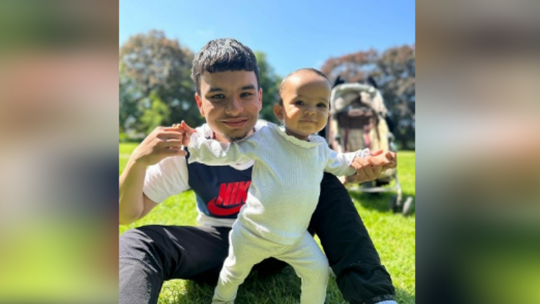 Mr Qureshi, wearing a white and blue Nike T-shirt with a Nike logo on it, sitting on grass with his baby daughter as she stands up and they smile at the camera