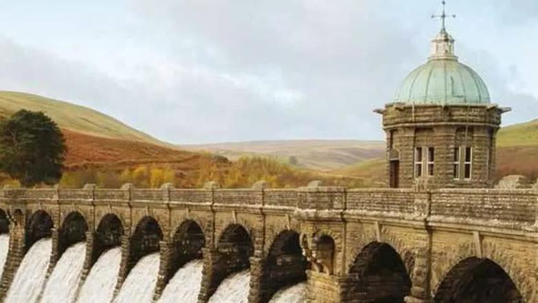 The Elan Valley Aqueduct, which consists of many stone arches which allow water to flow through, and a lead-roofed control building, against a backdrop of the Welsh hills