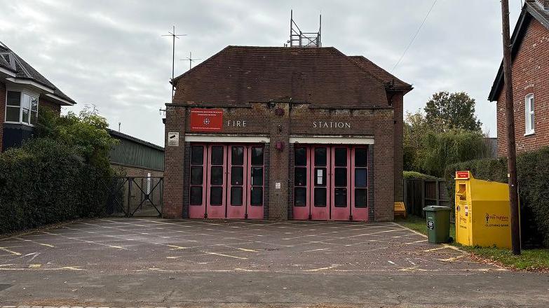 Single-storey brick-built fire station, with light red steel and glass doors across the whole of the front facade. There is a pitched roof with aerials mounted on it. There is a block-paved drive at the front criss-crossed with yellow paint. There is a yellow skip to the right for donated clothing, and houses on the left and right sides of the fire station site.