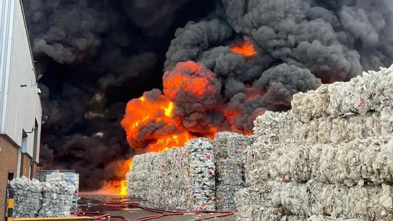 Bales of light-coloured material stacked in four levels. There are clouds of dark grey smoke with flame visible behind the bales of plastic. The smoke in the middle of the picture is almost black. There is a warehouse building to the left with a brick ground floor and white panelled upper floor.