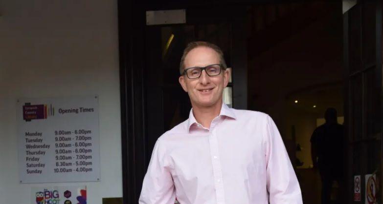 Bruce Leeke wearing a light pink shirt standing in a library. There is an opening-hours sign on his left.