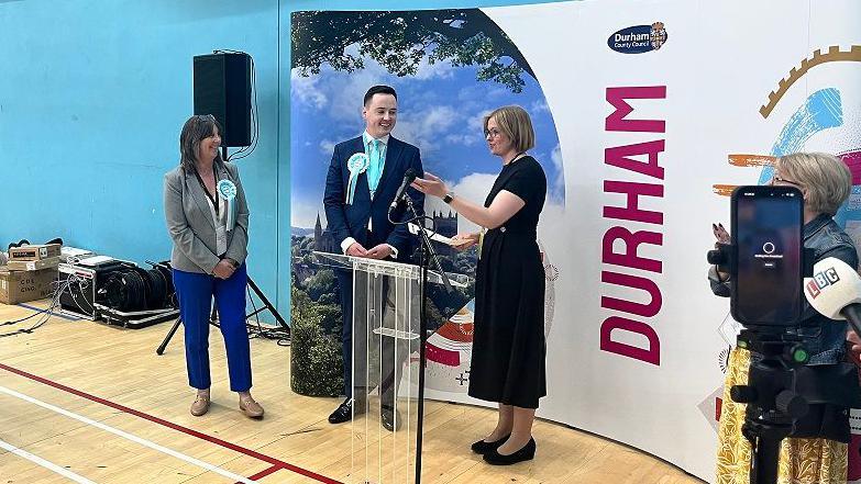 Darren Grimes, centre, on election night wearing a suit with a light blue tie and light blue and white Reform UK rosette. His is tall with dark hair. Karen Allison stands next to him. She had long dark hair and wears blue trousers. Journalists are next to them, as well as a woman who is announcing the election results