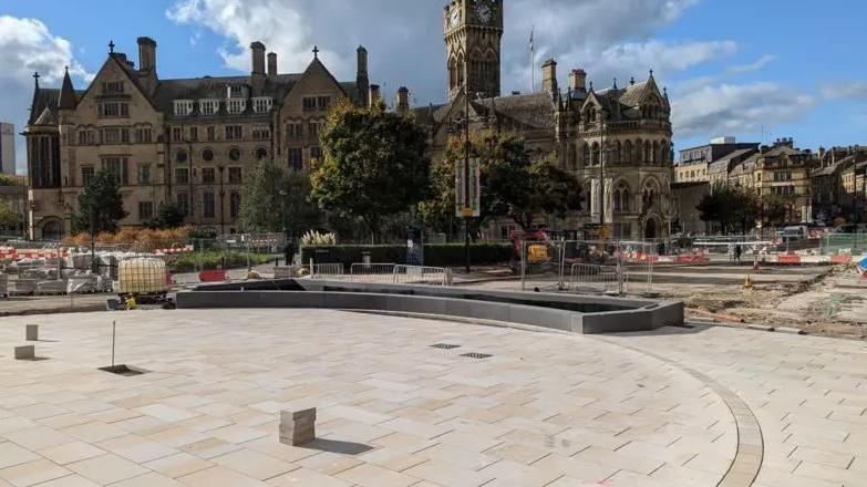 Paving work taking place in Bradford city centre, with fencing preventing pedestrians entering the construction site. Bradford City Hall is visible in the distance, along with large trees. 